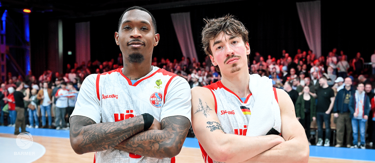 Zwei Basketball-Spieler der RheinStars Köln stehen mit verschränkten Armen vor Zuschauern.