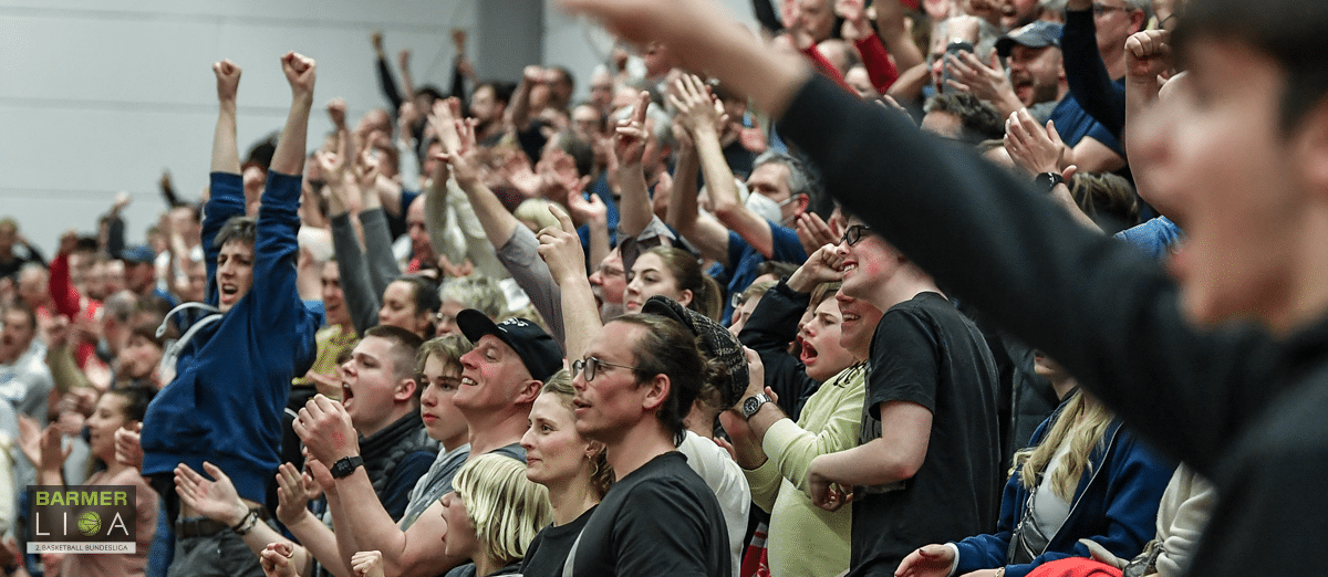 Enthusiastische Fans der RheinStars Köln jubeln mit erhobenen Armen in einer Sporthalle.