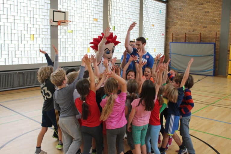 Kinder jubeln in einer Sporthalle um einen Maskottchen-Charakter mit roten Haaren herum.