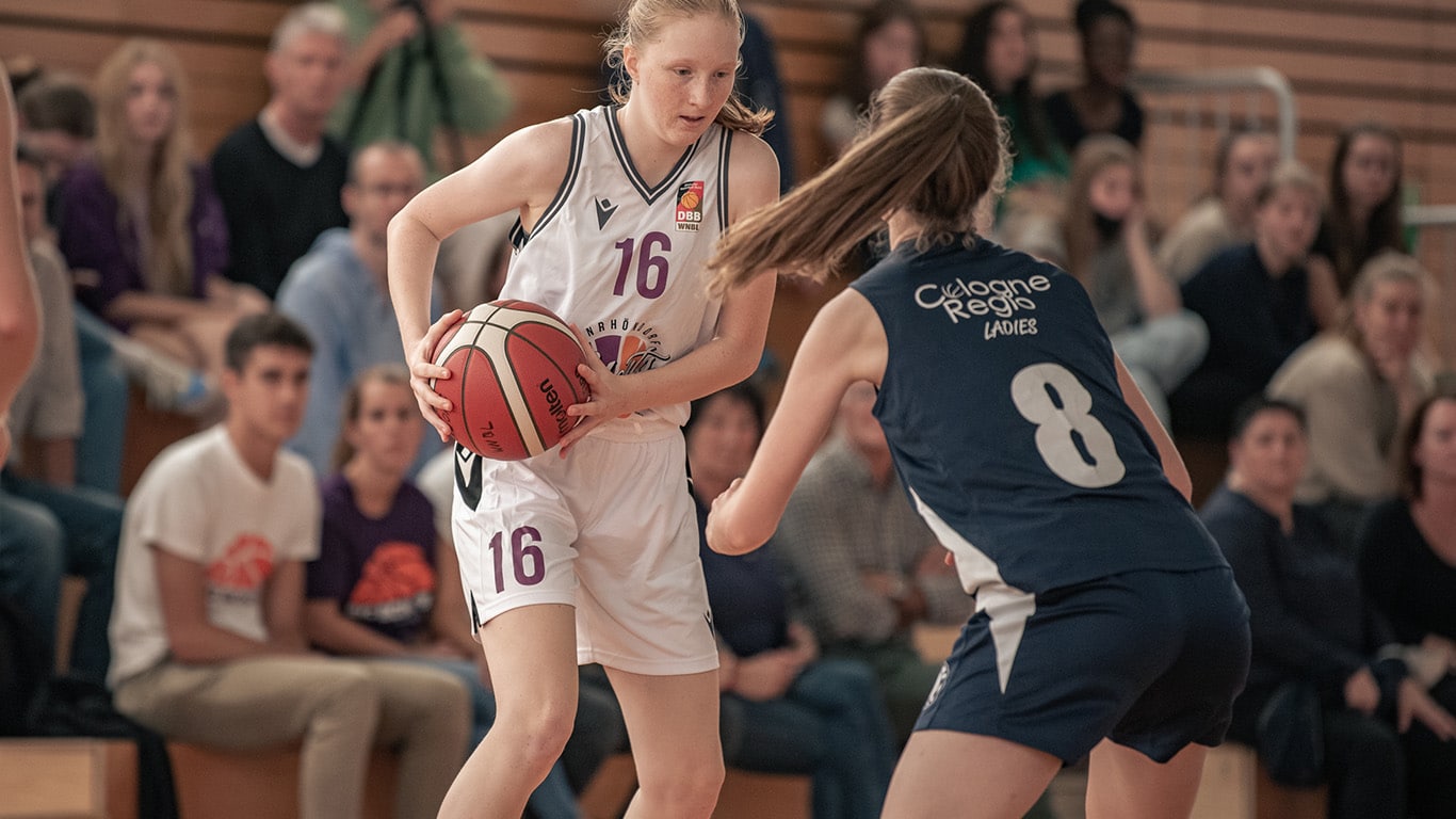Basketballspielerin der RheinStars Köln Nummer 16 mit Ball gegen Abwehrspielerin Nummer 8 in einer Sporthalle.