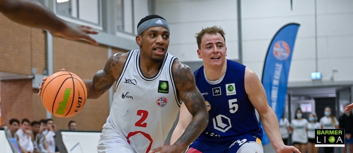 Basketballspiel der RheinStars Köln: Spieler in weißem Trikot mit Nummer 2 dribbelt gegen Gegner in blauem Trikot Nummer 5.