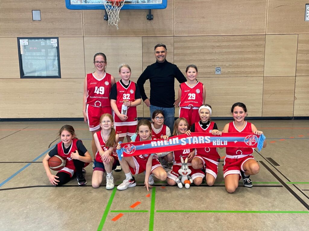 Mädchen-Basketballteam der RheinStars Köln posiert mit Schal in Sporthalle.