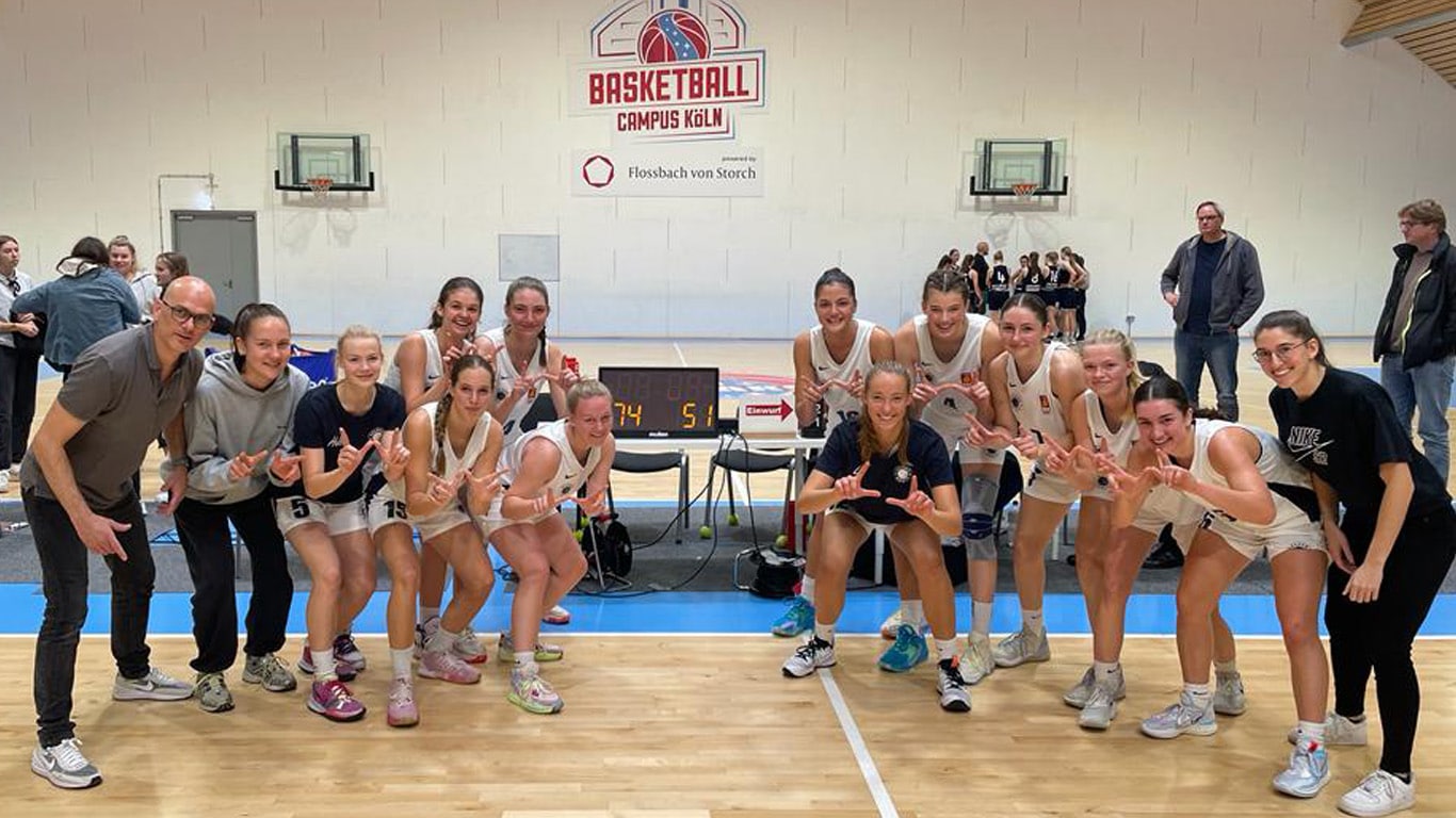 Frauenbasketballmannschaft der RheinStars Köln posiert lachend auf dem Spielfeld einer Sporthalle.