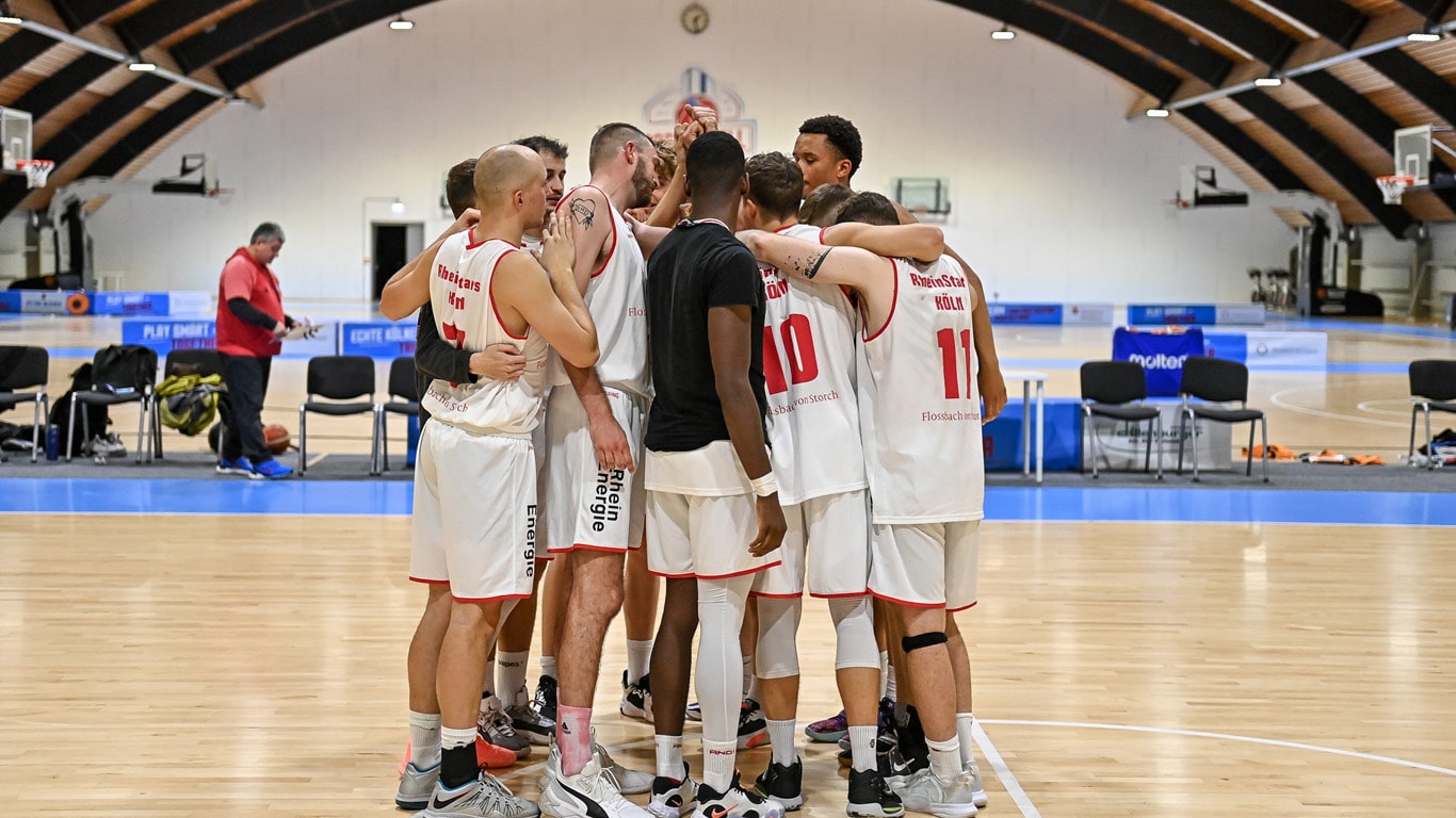 Basketballmannschaft der RheinStars Köln im Teamkreis auf dem Spielfeld einer Sporthalle.