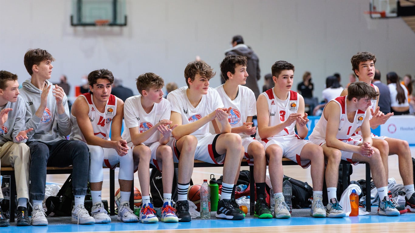 Jugendbasketballmannschaft der RheinStars Köln sitzt auf der Ersatzbank einer Sporthalle.