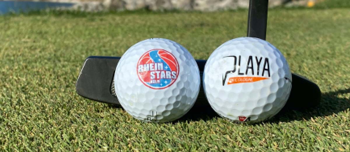 Zwei Golfbälle auf Rasen: links RheinStars Köln-Logo, rechts Playa-Schriftzug, daneben schwarzer Putter.