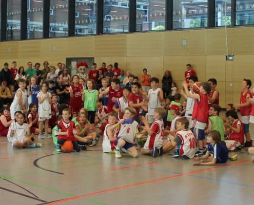Große Gruppe von Kindern der RheinStars Köln sitzt in einer Sporthalle um einen Basketball.